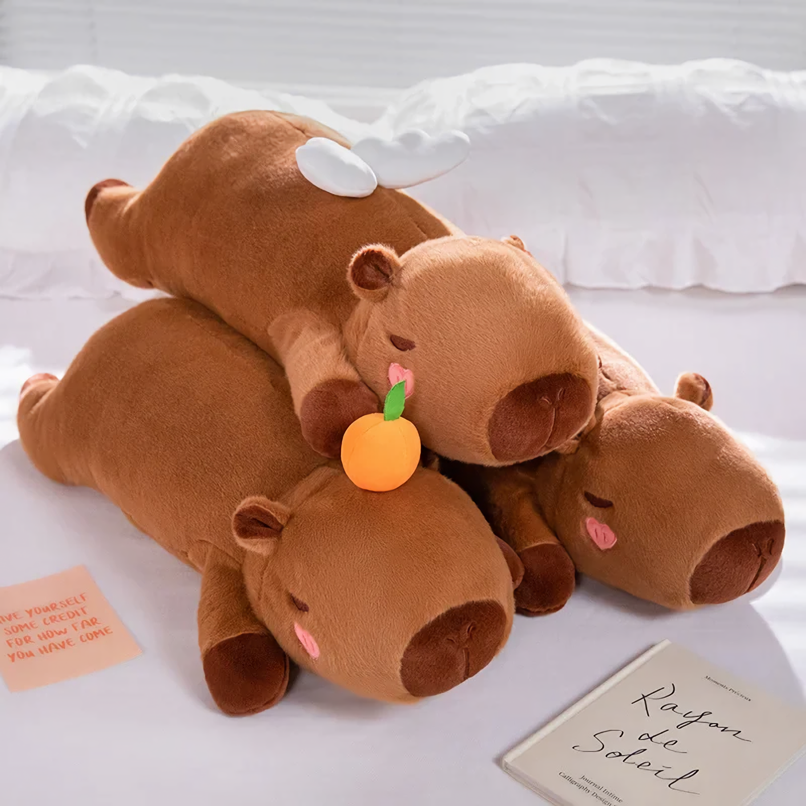 Three lying brown capybara plush pillows with white wings and an orange on a white bed