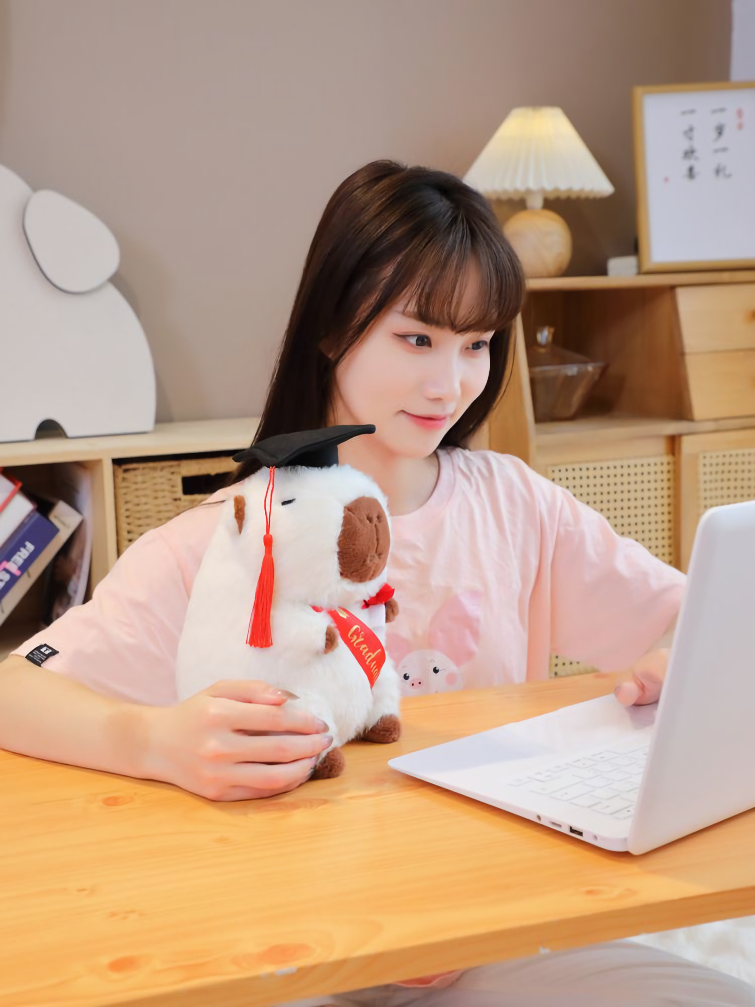 Woman at a desk with a laptop and a white graduation capybara plush