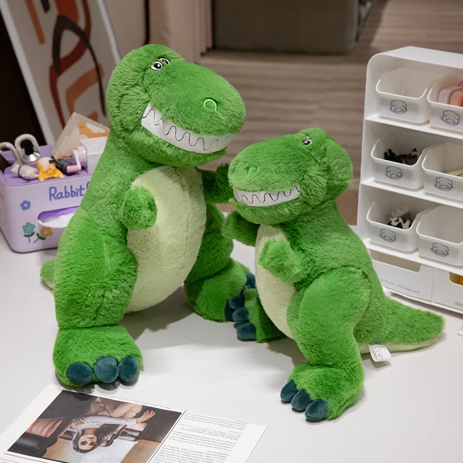 Two smiling green T-Rex dinosaur plush toys on a table, showing size difference; one larger, one smaller.
