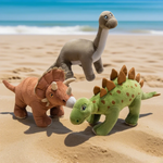 Gray Brontosaurus, brown Triceratops, and green Stegosaurus plushes on sandy beach with ocean in background.