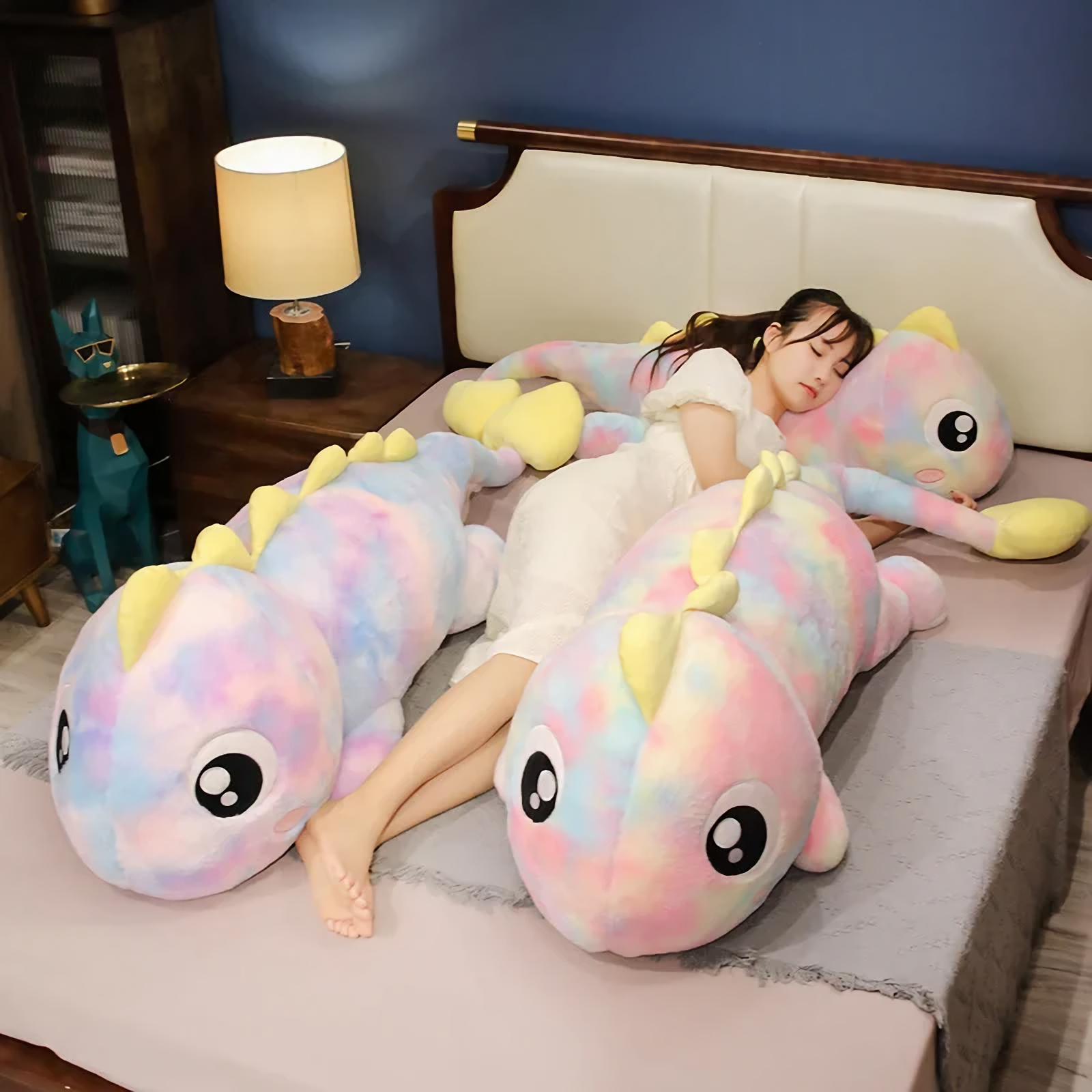 Woman lying on a bed beside two rainbow dinosaur plush body pillows