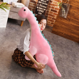Woman sitting on the floor holding a large pink Brachiosaurus dinosaur plush with blue spikes.