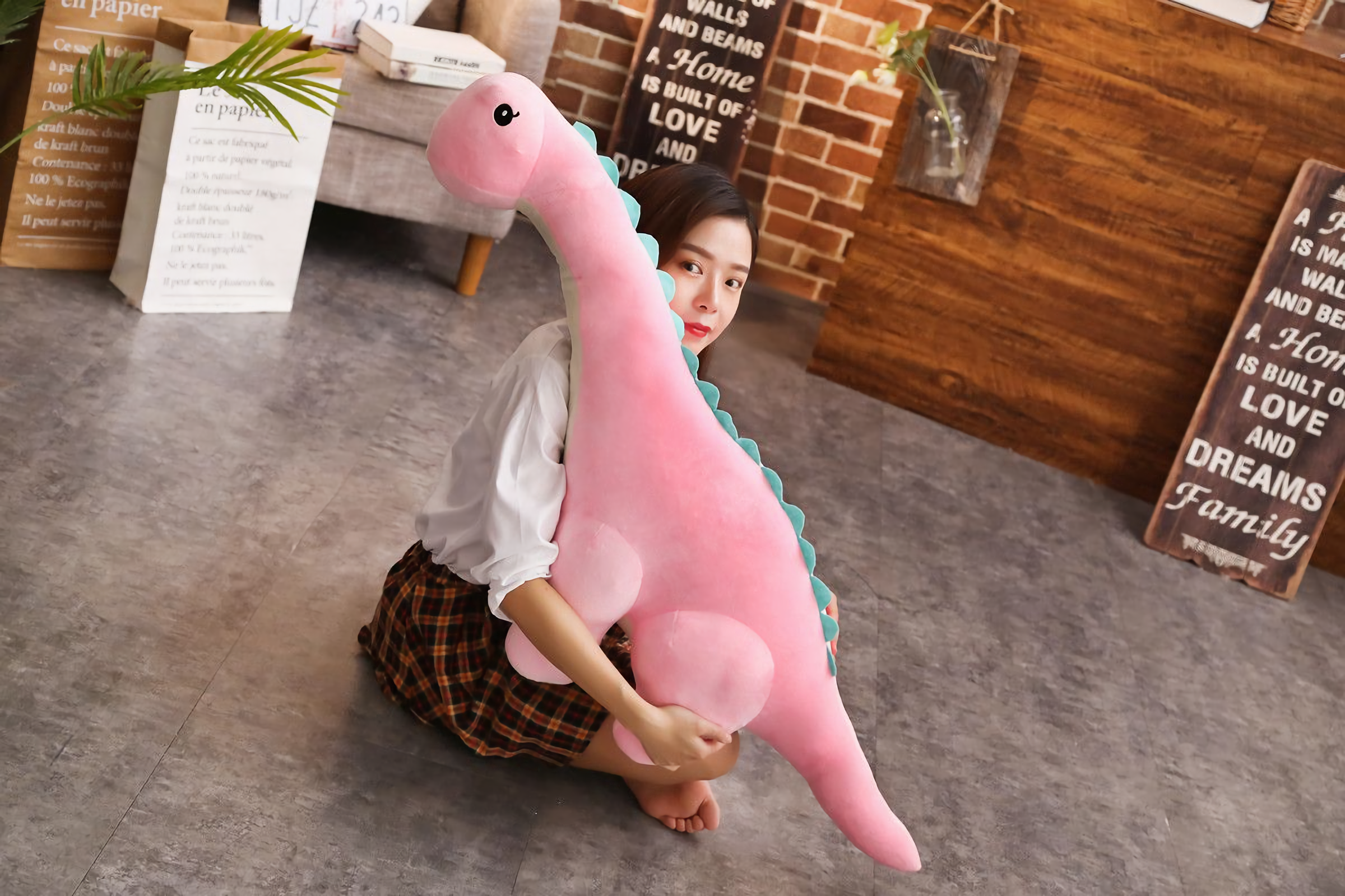 Woman sitting on the floor holding a large pink Brachiosaurus dinosaur plush with blue spikes.