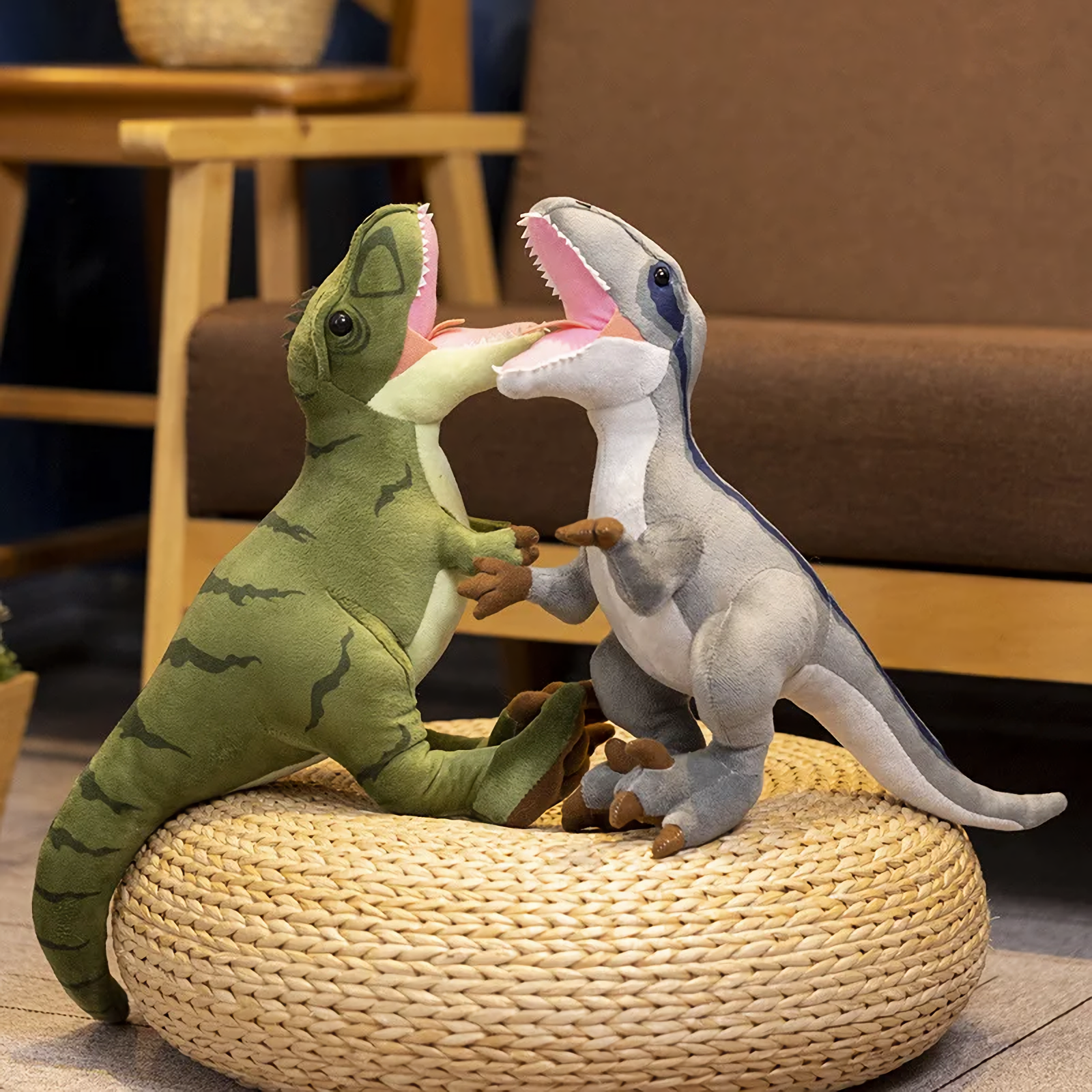 Green Tyrannosaurus Rex and gray Velociraptor plush toys on a woven pouf, facing each other with open mouths.