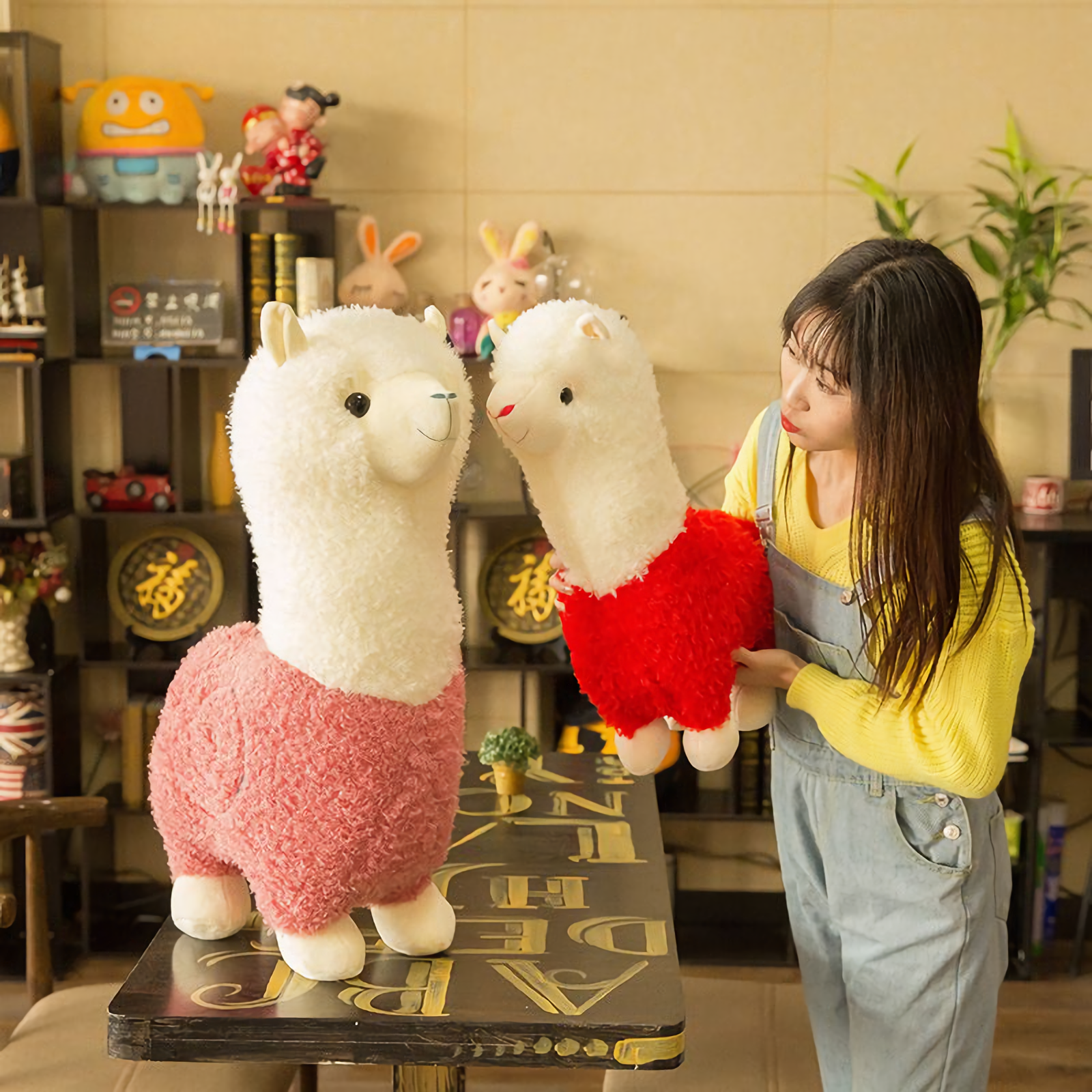 Woman hugging a red body alpaca plush beside a large pink body alpaca plush
