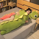 Woman hugging green dinosaur plush with yellow spikes on a bed. Pink dinosaur plush beside them.