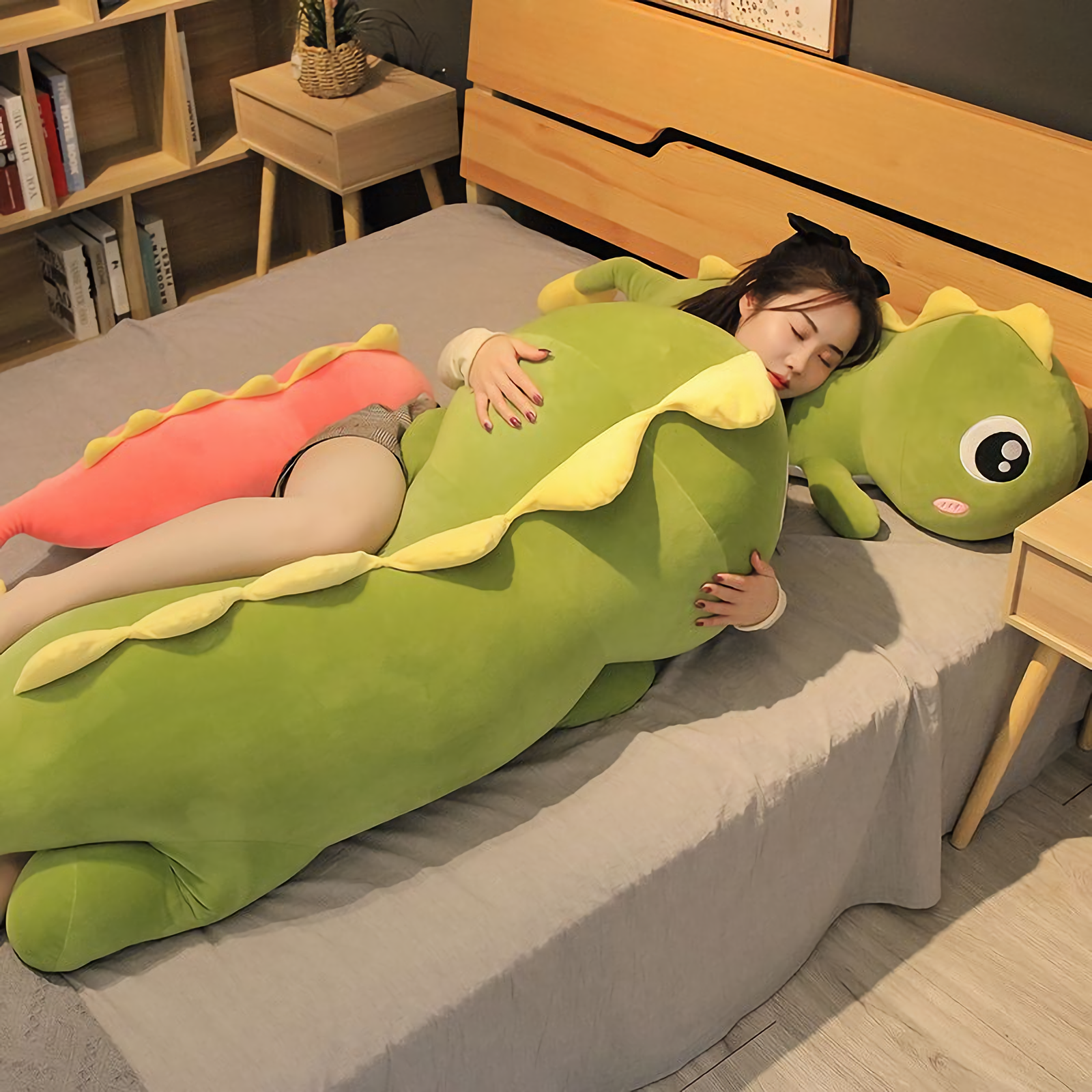 Woman hugging green dinosaur plush with yellow spikes on a bed. Pink dinosaur plush beside them.