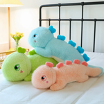 Three dinosaur plush pillows in green, pink, blue on a bed, soft heart-shaped spikes visible, sunny room.
