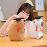 Woman resting her chin on a desk beside a brown and a white graduation capybara plush