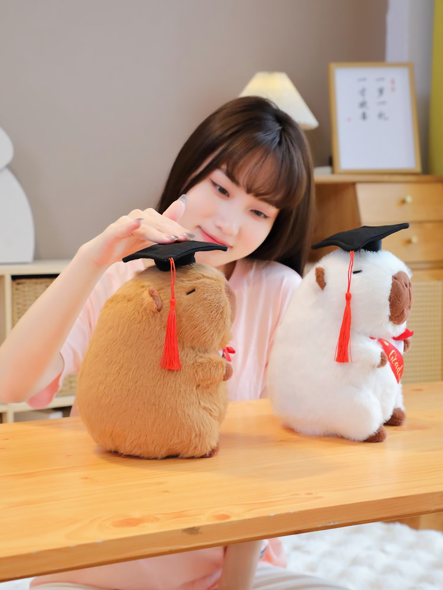 Woman resting her chin on a desk beside a brown and a white graduation capybara plush