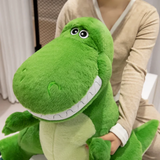 Woman holding a big friendly green T-Rex dinosaur plush with a wide toothy smile sitting on a bed.
