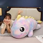Woman lying on her stomach smiling next to a blue and green rainbow dinosaur plush pillow