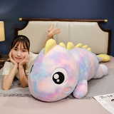 Woman lying on her stomach smiling next to a blue and green rainbow dinosaur plush pillow