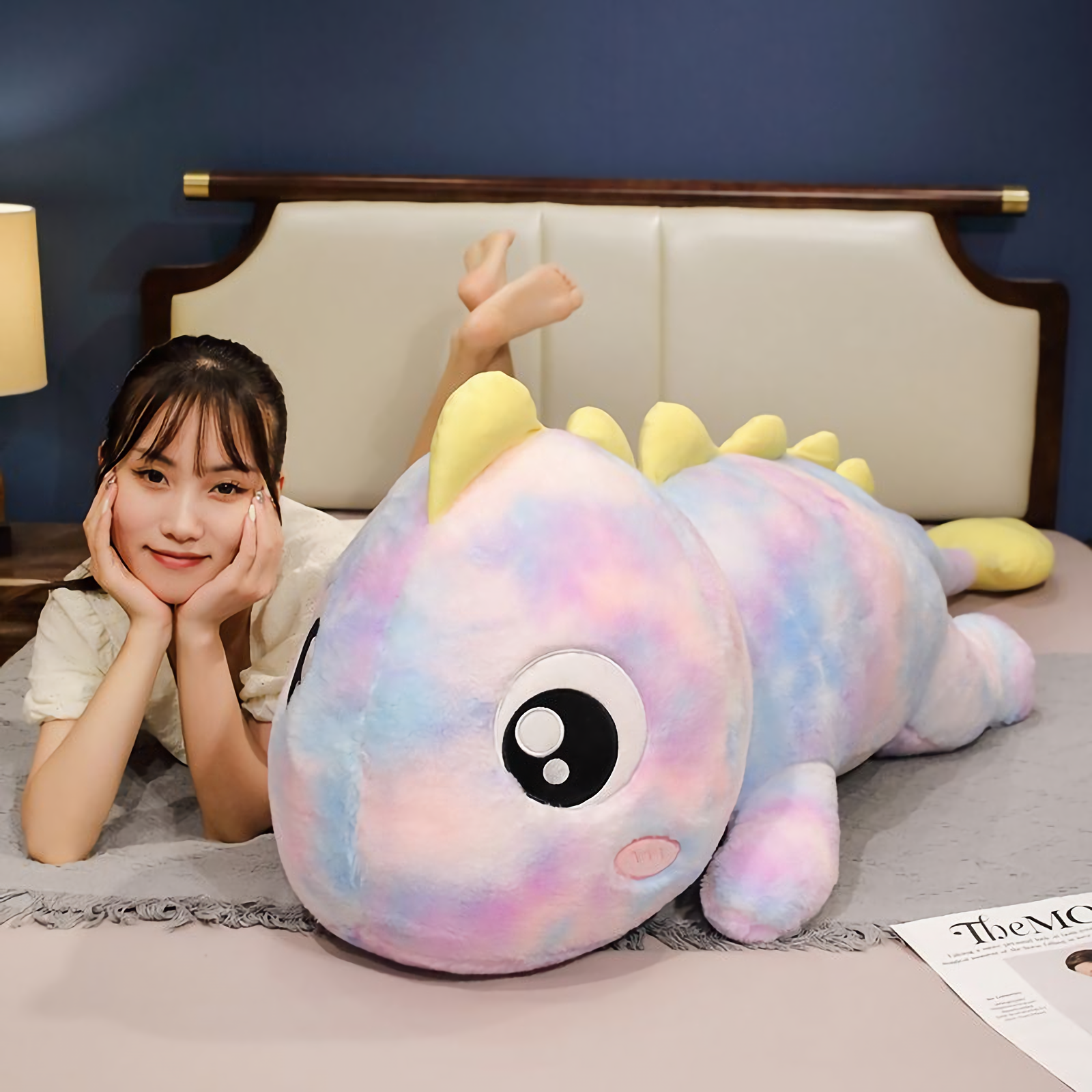 Woman lying on her stomach smiling next to a blue and green rainbow dinosaur plush pillow