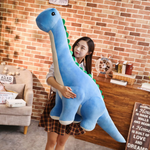 Woman holding large blue Brachiosaurus plush with green spikes and cream belly against a brick wall.
