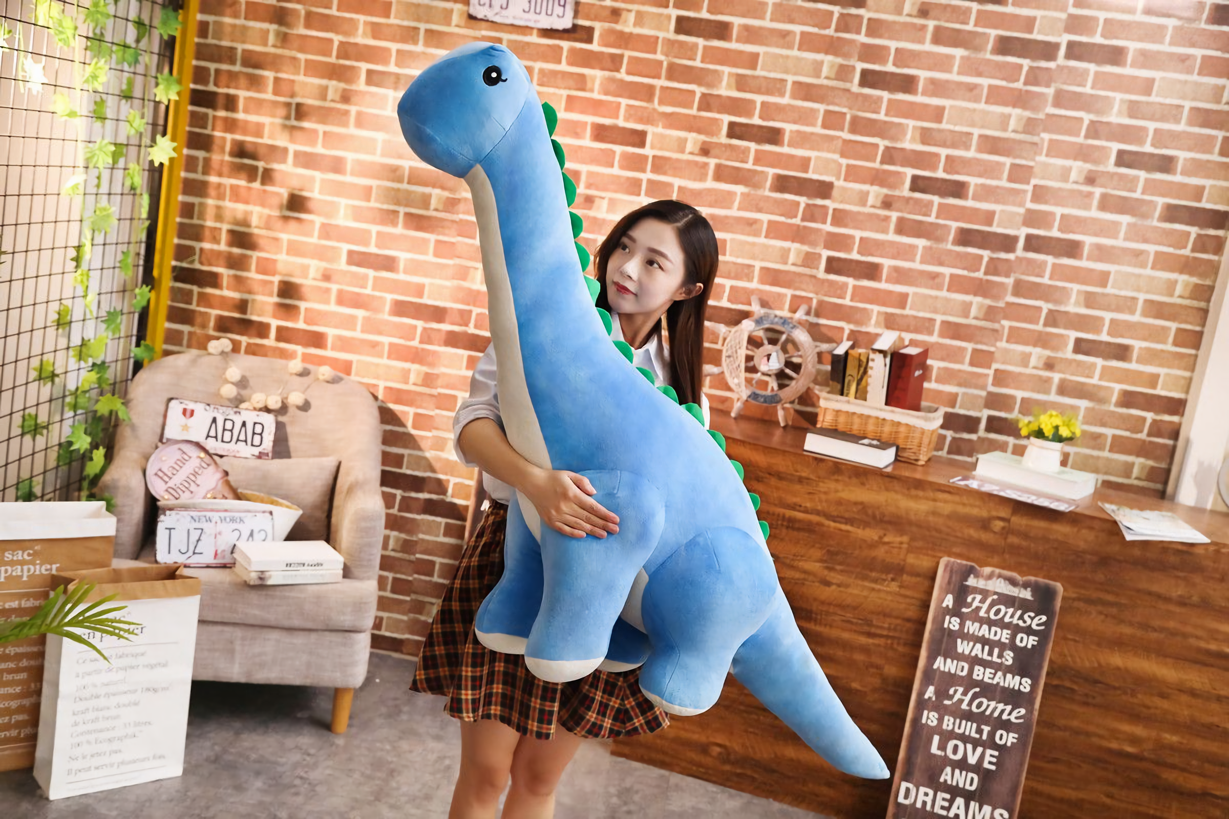 Woman holding large blue Brachiosaurus plush with green spikes and cream belly against a brick wall.
