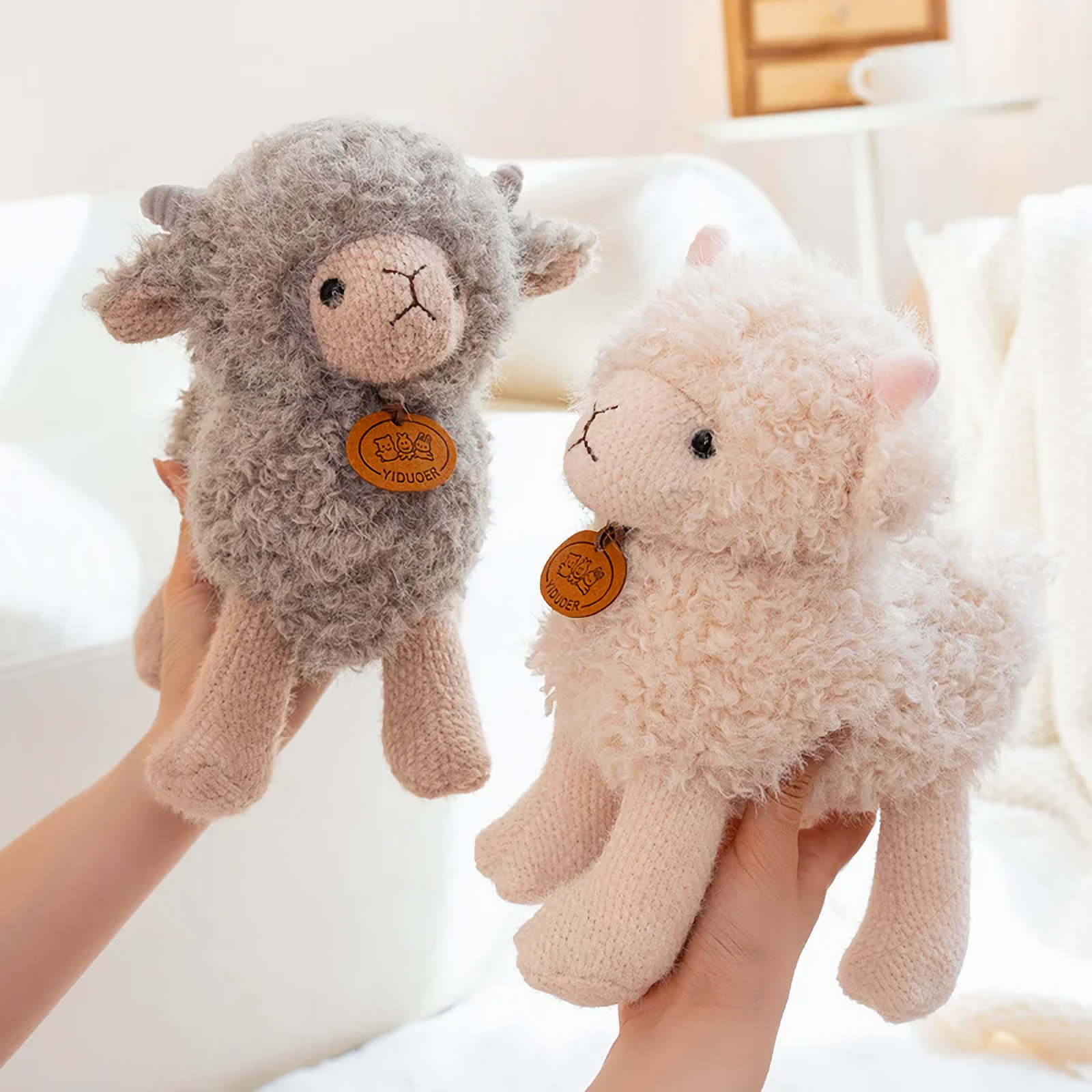 Hand holding a gray curly lamb plush beside a cream one on a bed