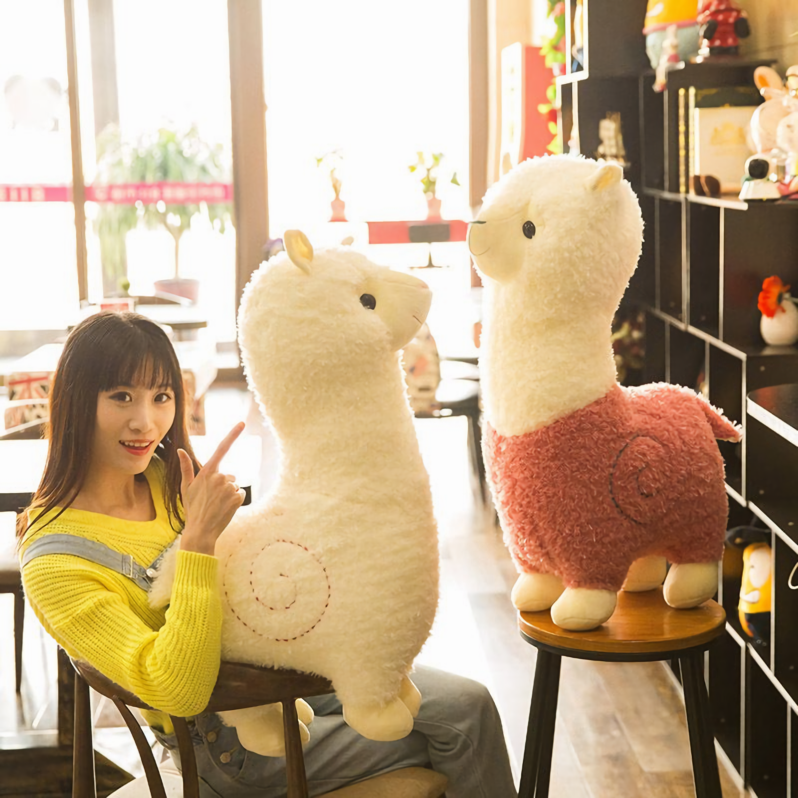 Woman sitting with a white alpaca plush beside a pink body alpaca plush