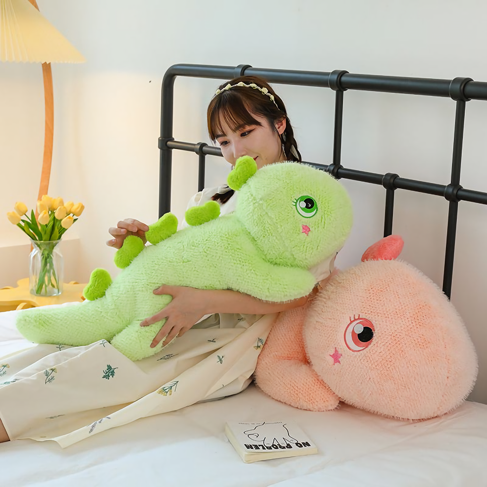 Woman holding green dinosaur plush pillow, pink plush beside, on a bed.