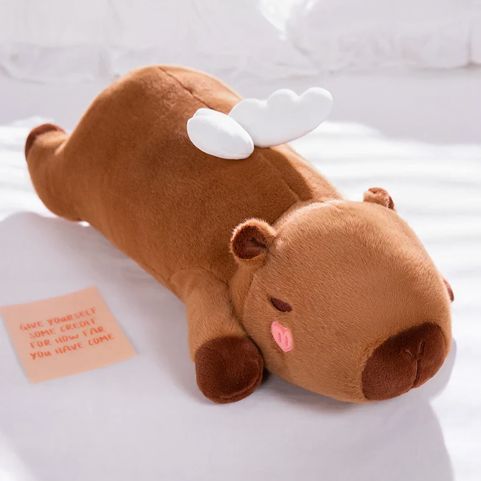 Lying brown capybara plush pillow with small white wings on a white bed
