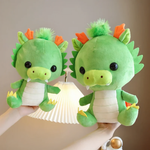 Two green dragon plushies with orange horns held up, one larger than the other, near a lamp.