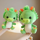 Two green dragon plushies with orange horns held up, one larger than the other, near a lamp.