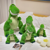 Five green T-Rex dinosaur plushies in different sizes on a bed with white bedding and a flower-shaped pillow.
