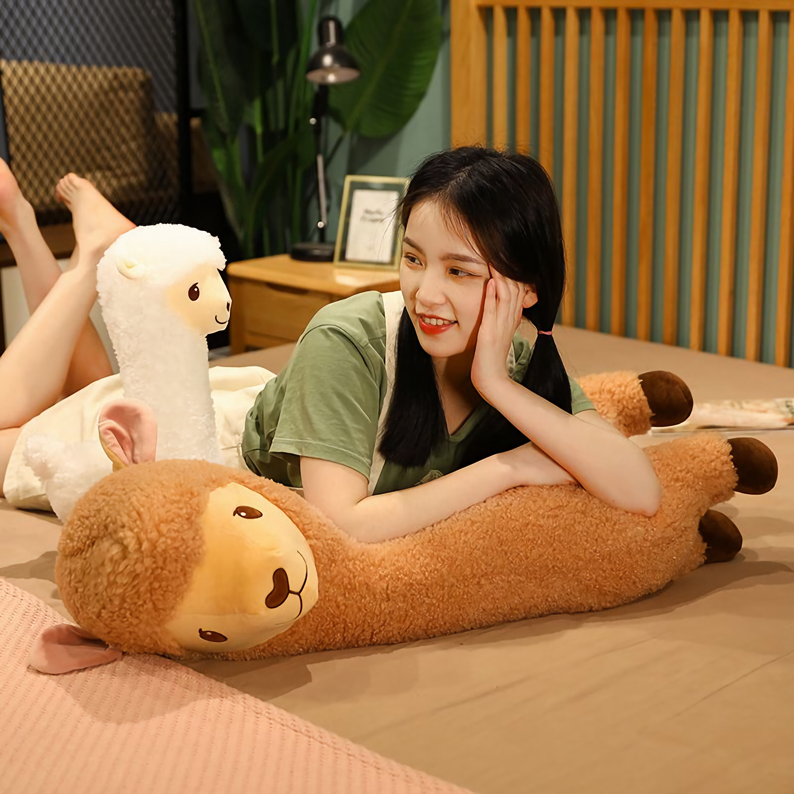 Woman lying on a bed beside a long brown alpaca plush and a white one