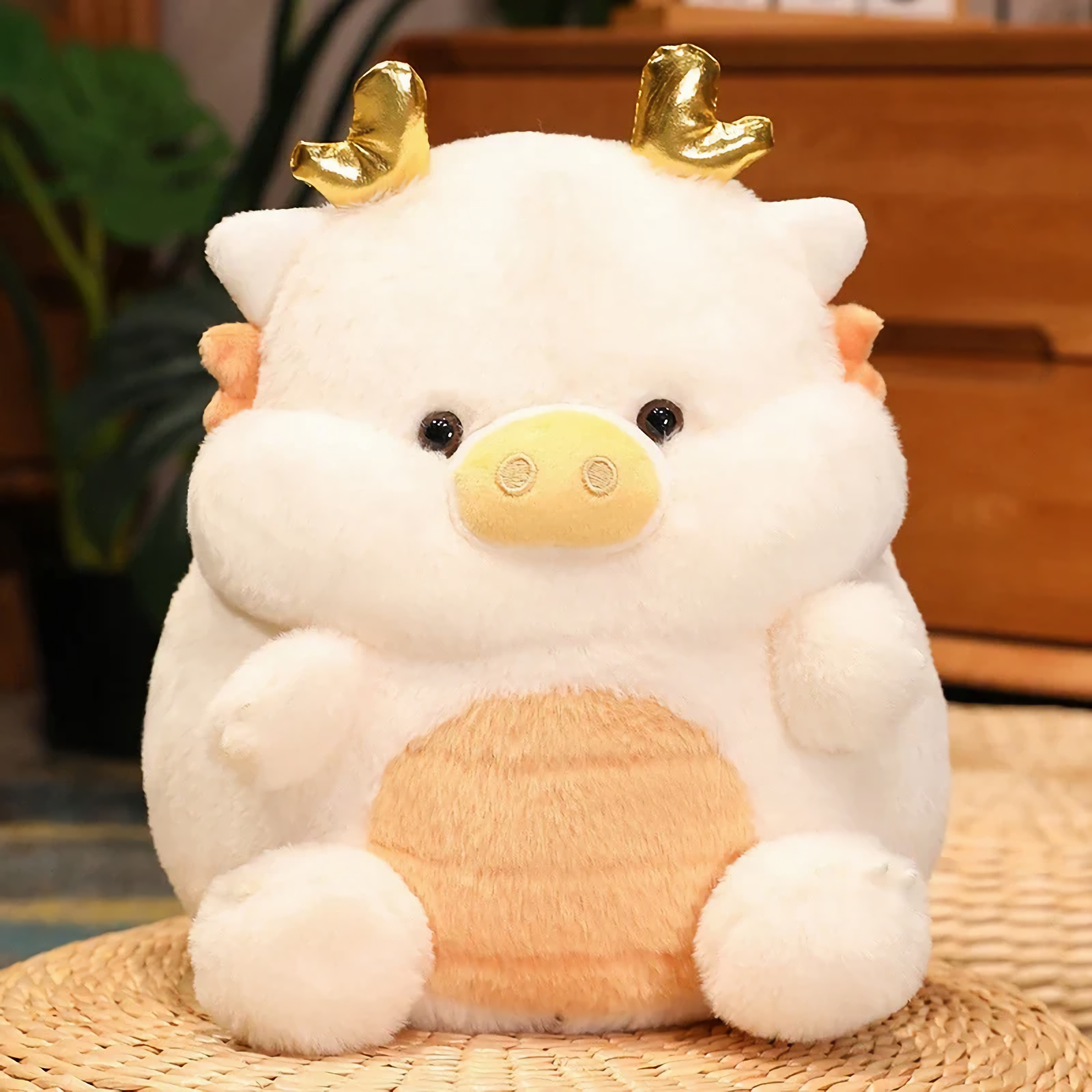 White pig-dragon plush cushion with golden horns on a wicker mat, showing its cream belly in a cozy indoor setting.