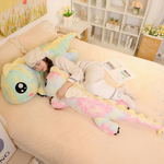 Woman lying on a bed beside a long rainbow dragon plush with a green face
