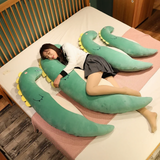 Woman hugging a curved green dinosaur plush pillow with more curved plush pillows on the bed