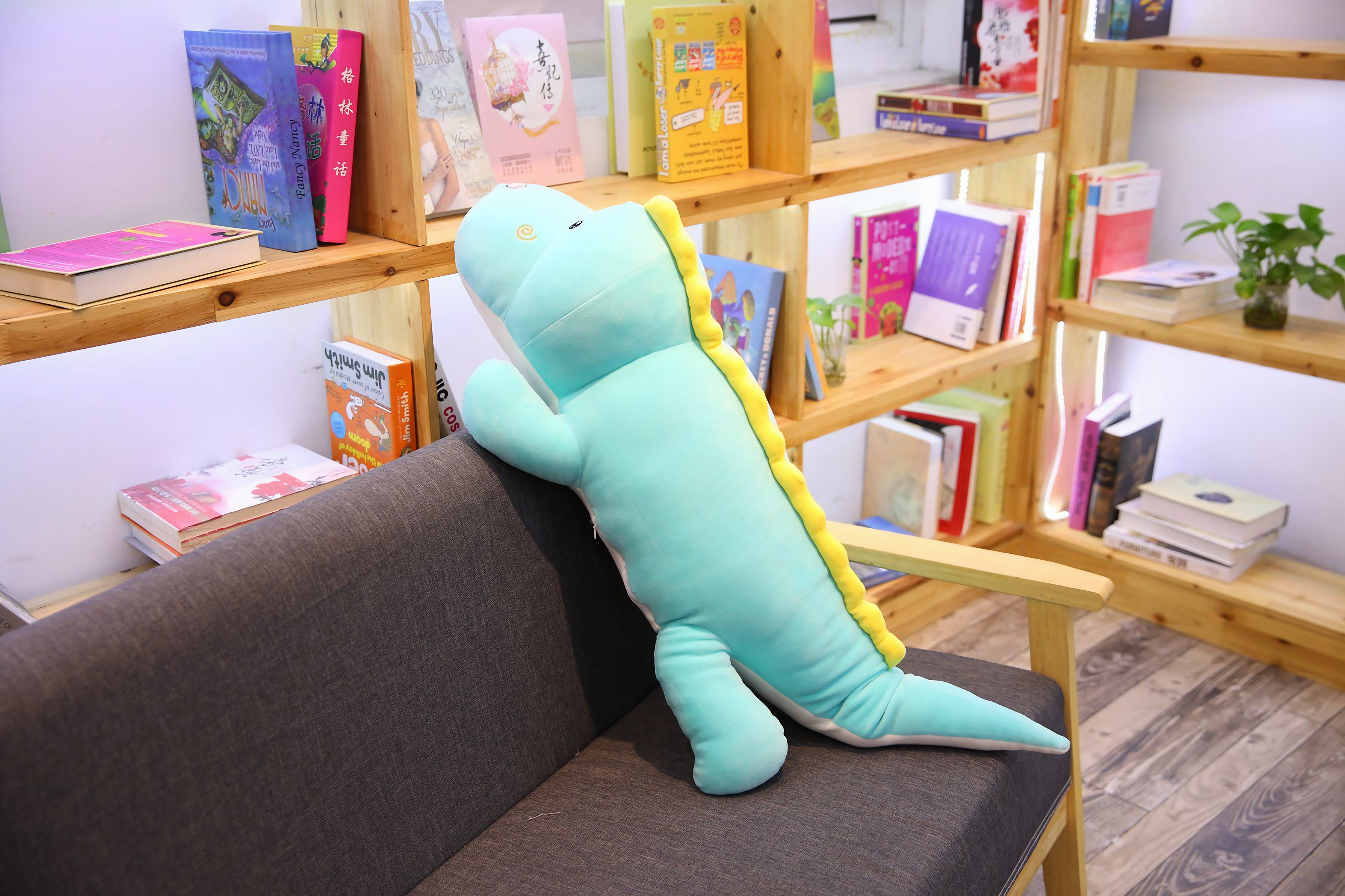 Long blue dinosaur plush body pillow leaning against a dark sofa in a bright room