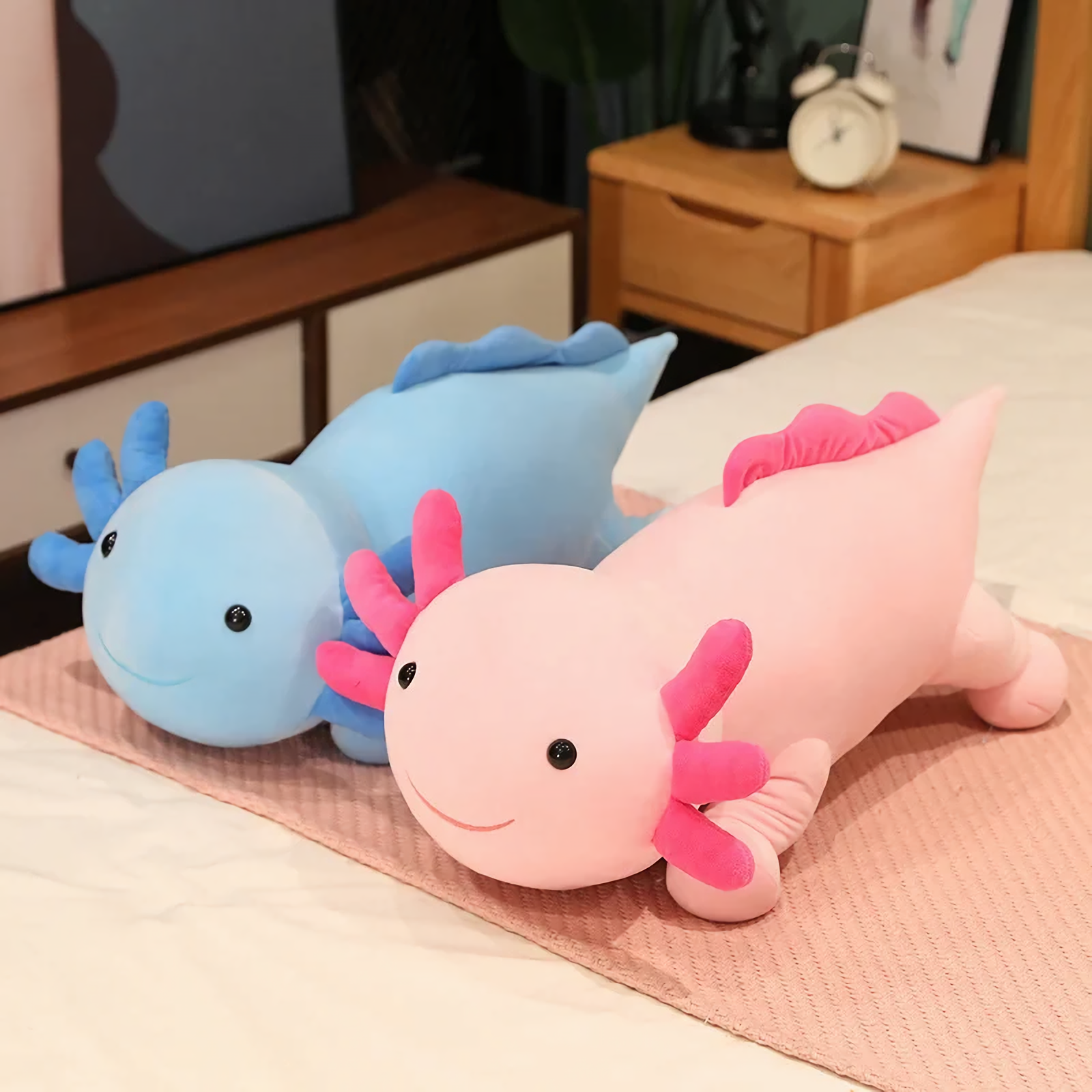 Blue axolotl plush and a pink axolotl plush lying on a pink blanket on a bed