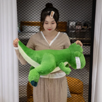 Woman holding smiling green T-Rex dinosaur plush horizontally, showing pale green belly and dark teal claws.