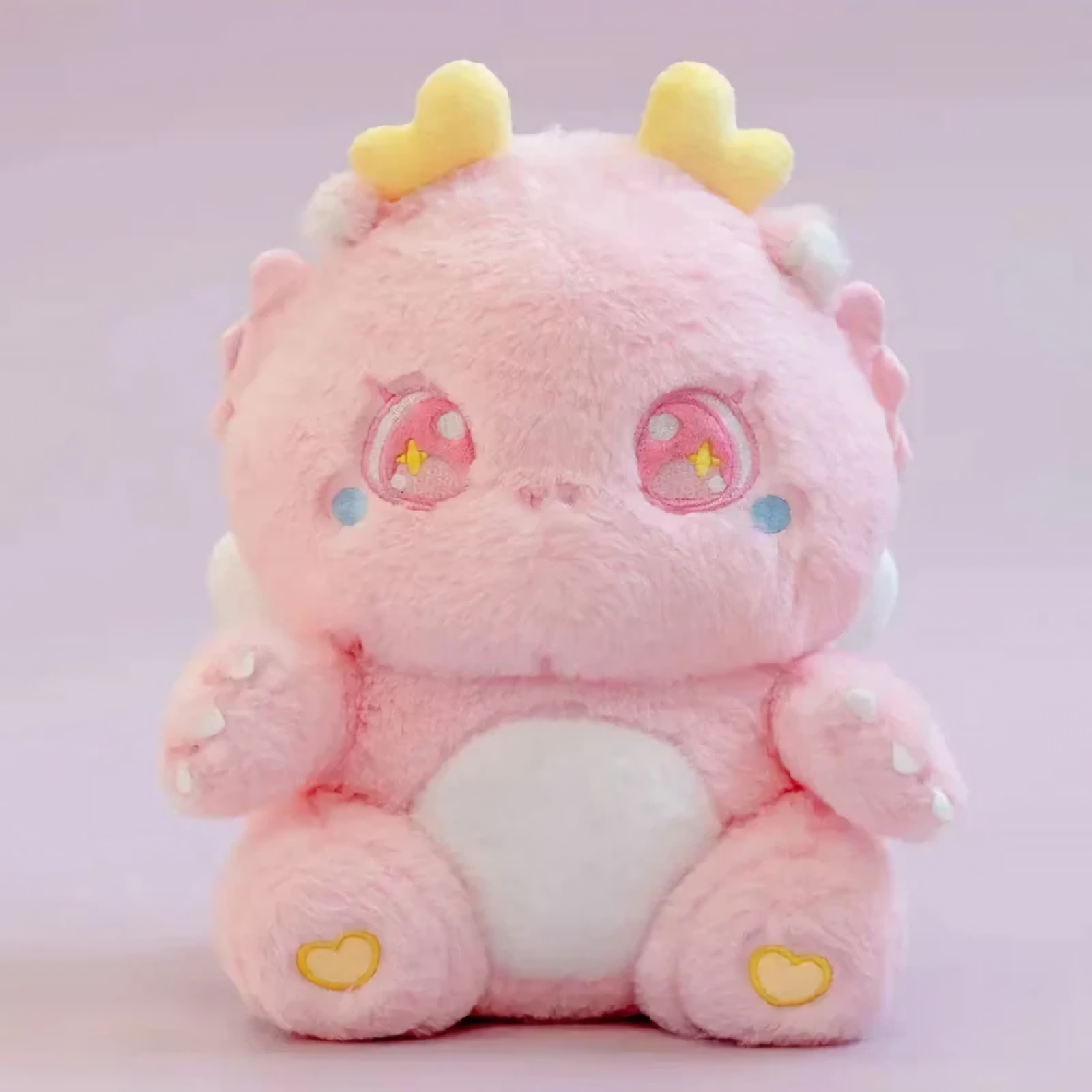 Pink fluffy baby dragon plush with sparkly star eyes and golden horns, sitting on a pink background.