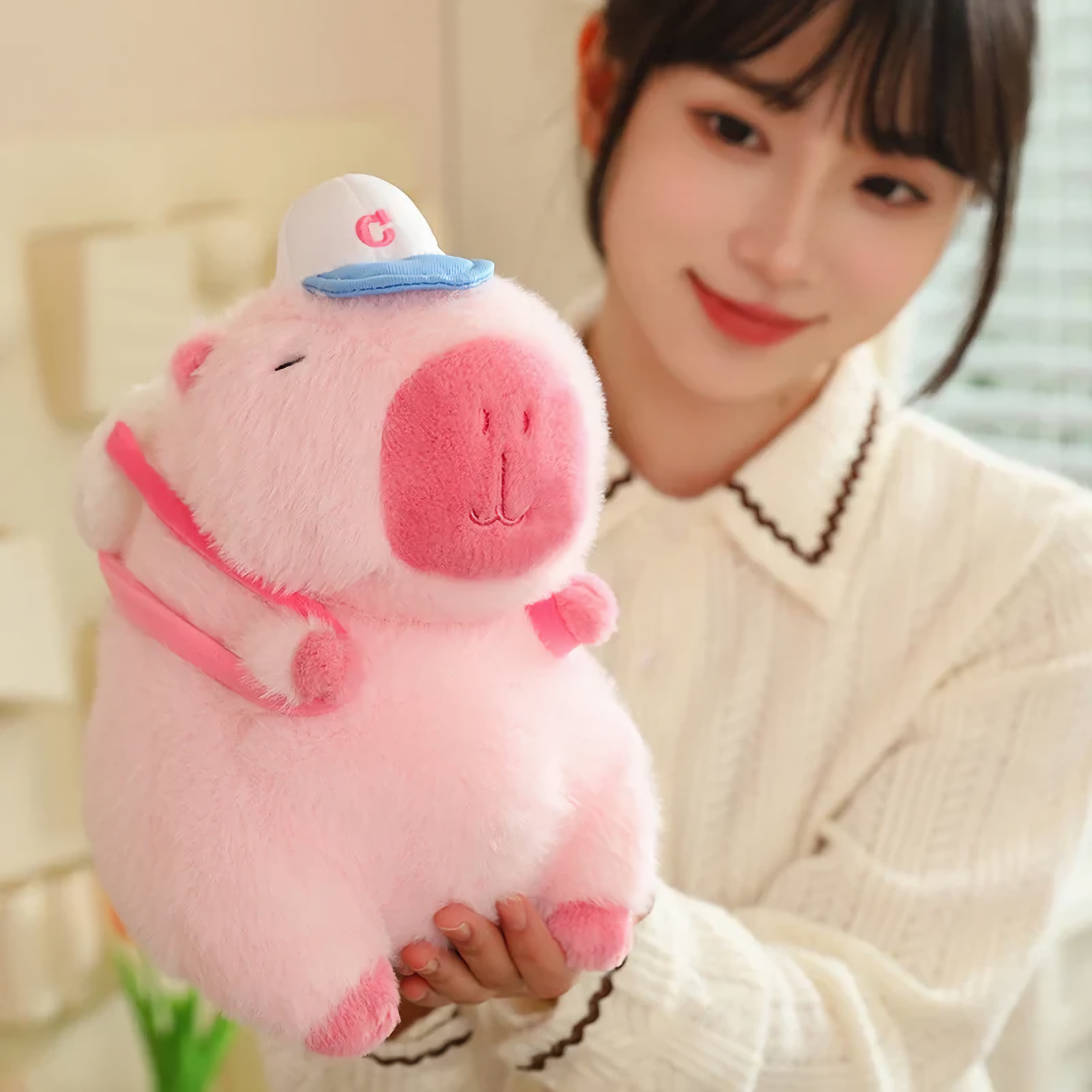 Woman smiling while holding a pink capybara plush with a white cap
