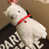 Hand holding a white alpaca plush by the ear to show its red bell collar