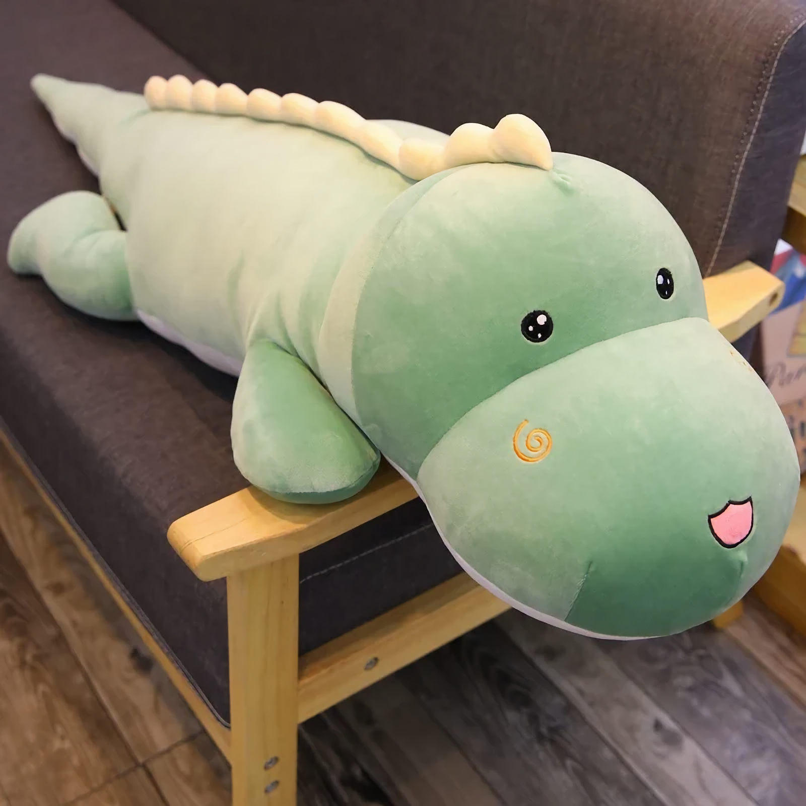 Green dinosaur plush body pillow lying on a dark sofa showing the cream back ridge