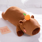 Lying brown capybara plush pillow with a small orange on its head on a white bed