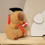 Brown graduation capybara plush with a black cap, red tassel and diploma on a shelf