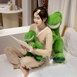 Woman reading with two green T-Rex plush toys, one on her lap and one behind her, on a bed.