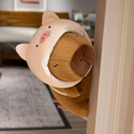 Capybara plush in a pink pig hood peeking around a door frame