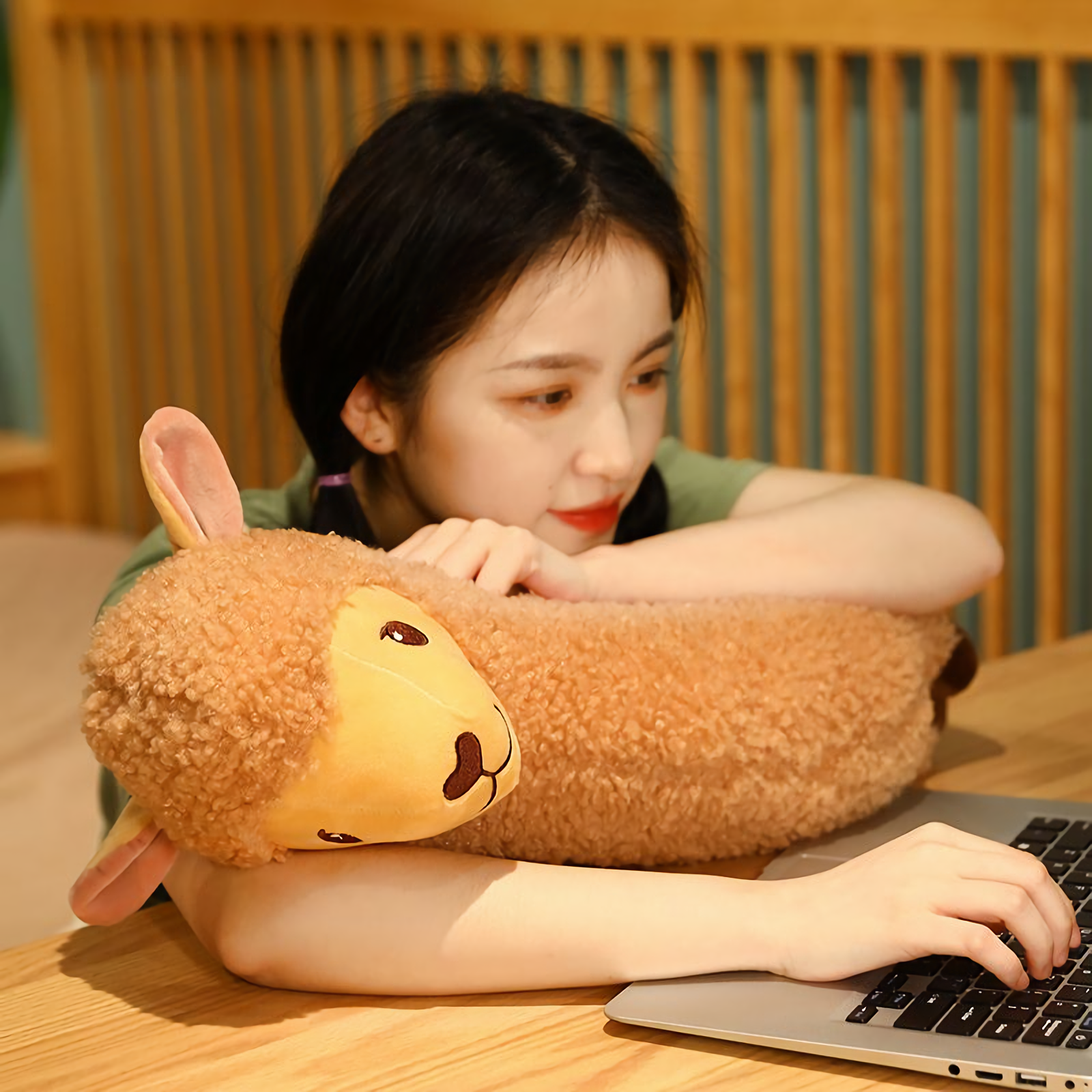 Woman resting her head on a long brown alpaca plush beside a laptop