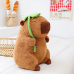 Sitting capybara plush holding a green lotus leaf on a white bed