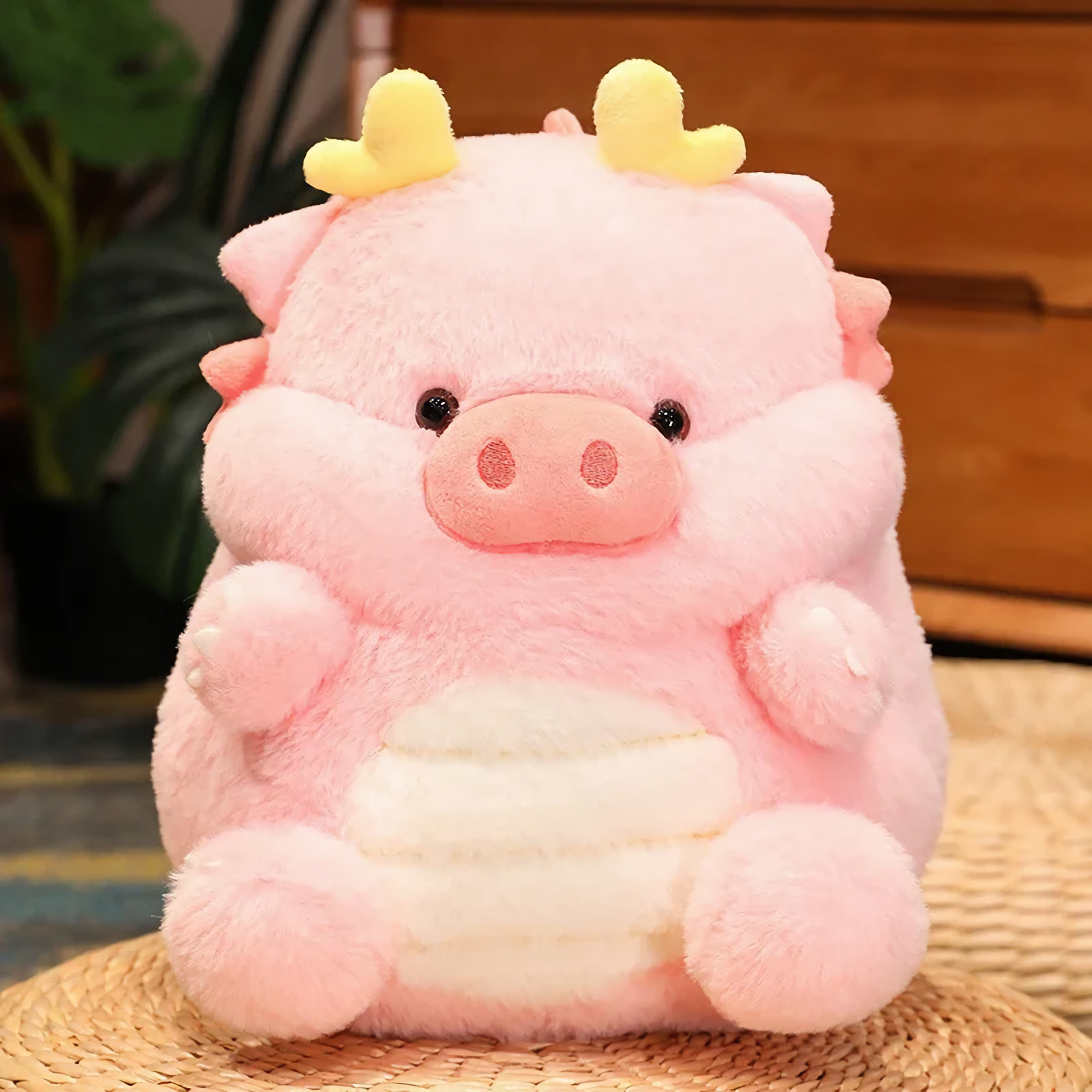 Pink pig-dragon plush cushion with cream belly on woven mat, close-up of face and tiny horns.