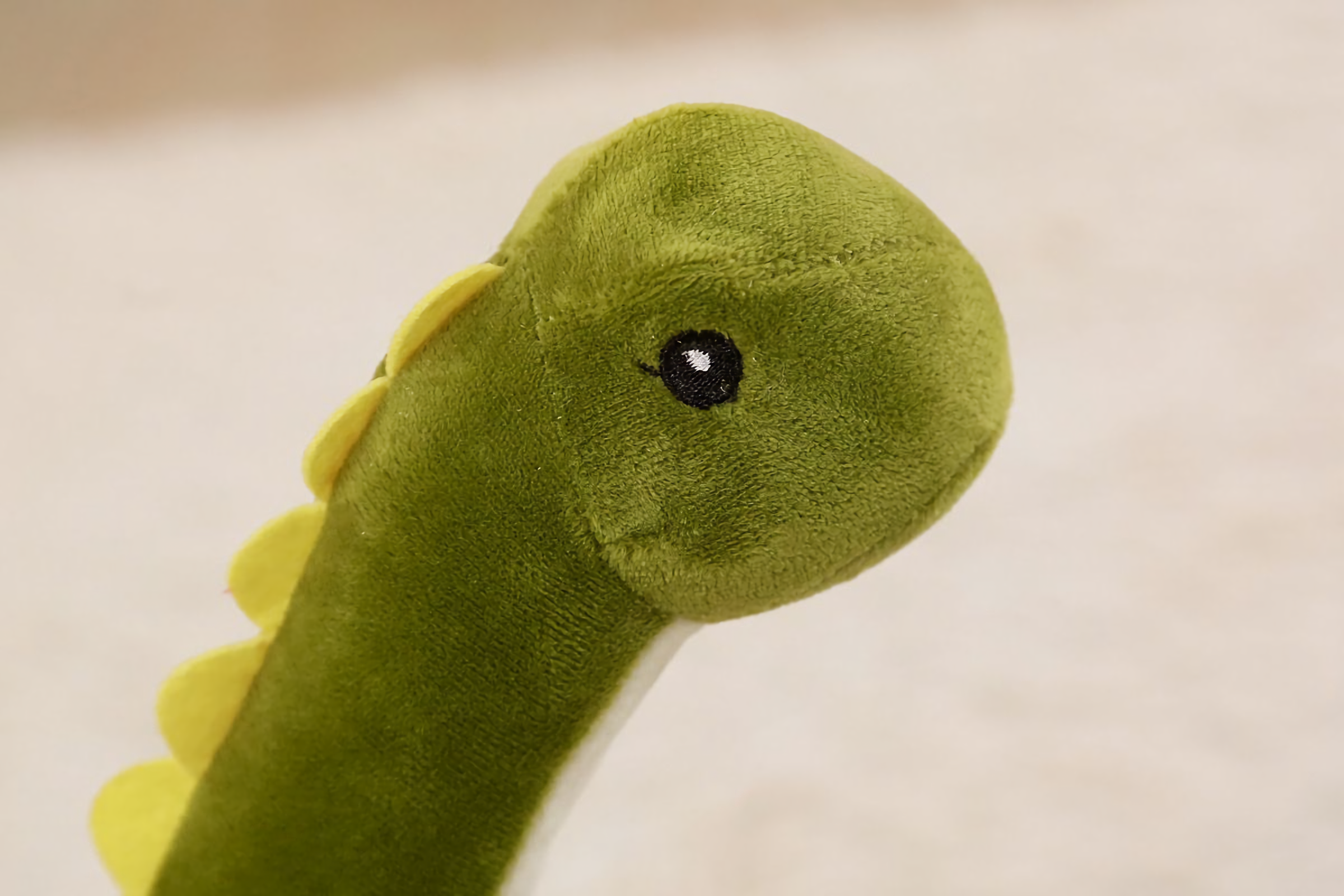 Close-up of green Brachiosaurus plush face with cream belly and yellow spikes, against a beige background.