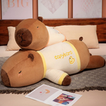 Two lying capybara plush pillows in cream shirts stacked on a bed