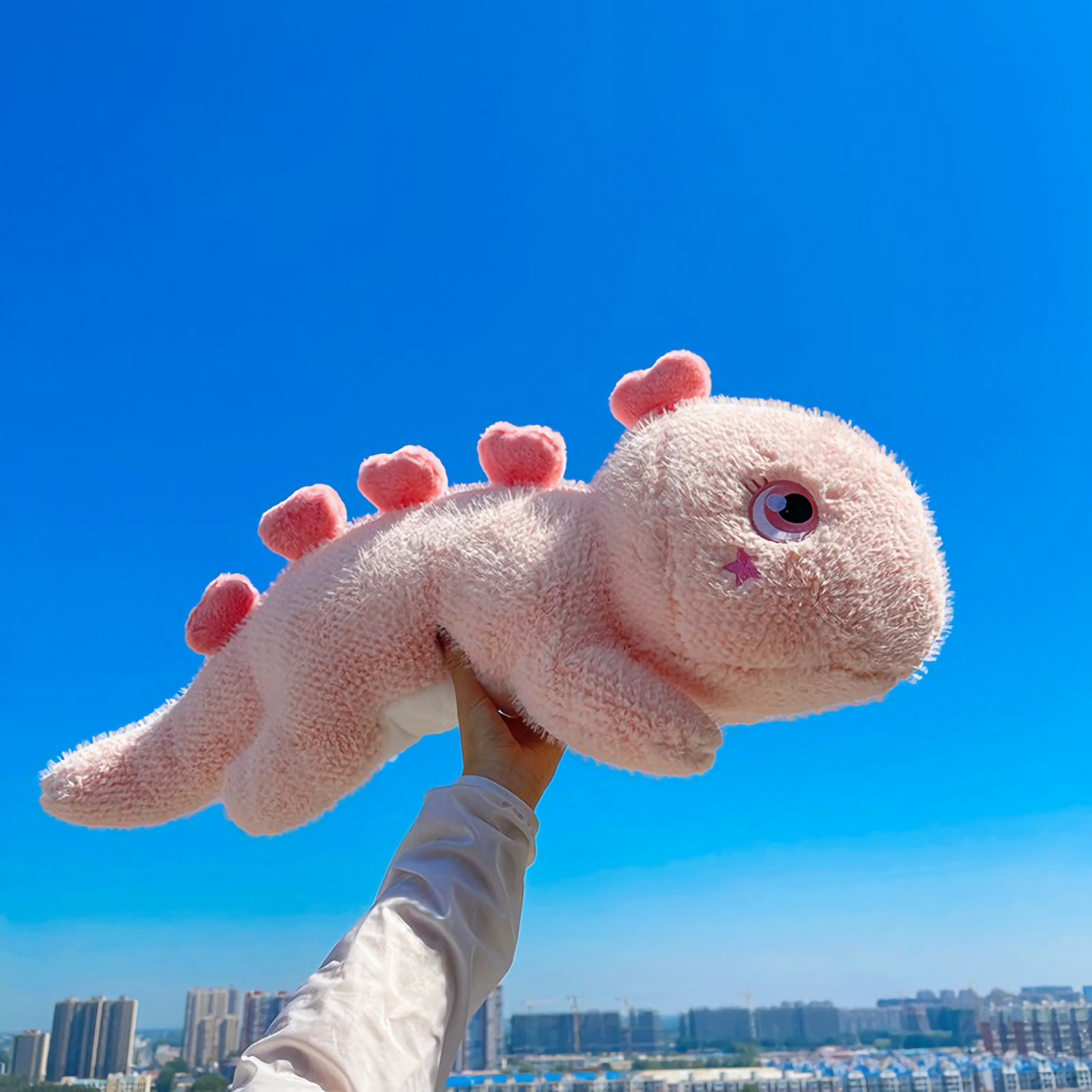 A pink dinosaur plush pillow with heart-shaped spikes held up against a clear blue sky.