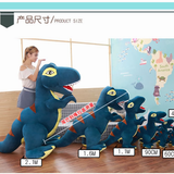 Woman with 2.1m blue and green T-Rex plush beside smaller plush sizes, in a room with a world map wall.