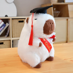 White graduation capybara plush with a black cap, red tassel and Graduate sash on a table